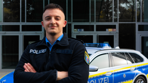 Ein Polizist in Uniform steht mit verschränkten Armen vor einem Streifenwagen und lächelt in die Kamera.