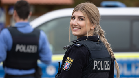 Eine Polizistin in Uniform ist von hinten zu sehen und lächelt über die Schulter in die Kamera. Im Hintergrund verschwommen ein weiterer Kollegen sowie ein Streifenwagen