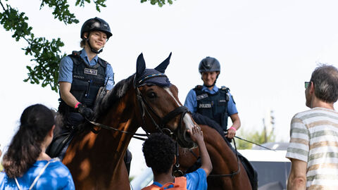 Zwei Polizistinnen sitzen auf Dienstpferden. Ein Kind streichelt den Kopf eines Pferdes
