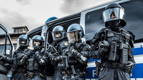 Fünf uniformierte Polizeibeamte stehen nebeneinander, ausgestattet mit Schutzwesten, Armschutz und Helmen. Hinter ihnen ist ein großer Polizei-Gruppenwagen geparkt.