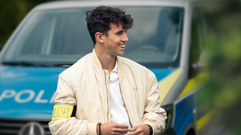 Ein Kriminalpolizist mit gelber Polizei-Armbinde steht lächelnd vor einem Polizeifahrzeug