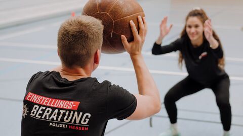 In einer Sporthalle trainieren zwei Personen: Eine Person hält in Kniebeugeposition einen Medizinball, eine andere steht gegenüber in Fangbereitschaft.