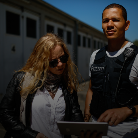 Kriminaloberkommissar in Schutzweste zeigt auf ein iPad, das eine Kriminaloberkommissarin in den Händen hält und liest.